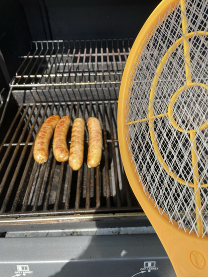 Bbq grill with 4 fennel sausages cooking. A yellow electric fly swatter that looks more like a mini tennis racket in the foreground