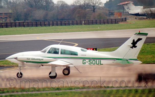 Side view of a twin engined light aircraft taxiing from right to left along a reddish-grey taxiway.
The plane is mostly white, with a green and black stripe running along the body, with the green registration "G-SOUL" on the lower rear fuselage.
There are green "Air Atlantique" titles on the wing-tip fuel tanks, and a black shadow of a stooping eagle on the tail.
Green grass lines the taxiway in the foreground, with more grass lining both sides of the runway in the background.
A black wooden fence stretches across the background from the left, with the tail and rear fuselage of a rather forlorn 3 engined jet airliner coming in to view of the right.
Trees in the distance meet bright but hazy sky.