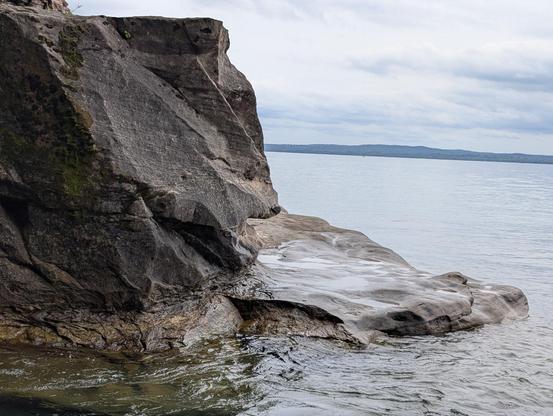 You can see the small hills in the distance across the water beyond this rock formation in front of you. The water is very clear but the surface is still reflective. A sort of flat-ish smoothly textured rock surface forms a platform just above the waters surface. Soft divots pool with water that's splashed up from the lake. A taller formation juts above to the left with sharp-ish but smooth looking faces and edges. Moss and lichen take advantage of the texture to make a home and you can see some plant matter atop this boulder as well.