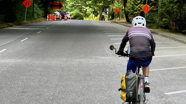 Single cyclist on a two-lane paved road approaches a construction zone beginning on the crest of a hill.