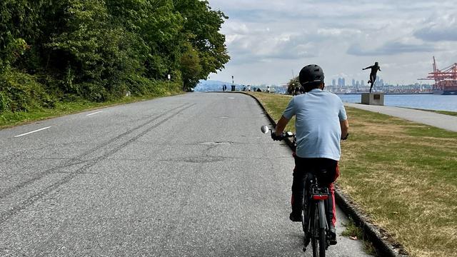 Single cyclist on a two-lane paved road approaches a statue of Harry Jerome on the right side of the road.