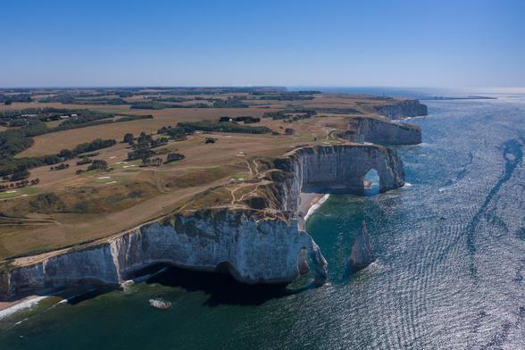 Aerial view of rugged white cliffs along a coast, with green fields and a shimmering ocean below. The landscape features natural arches and rocky formations near the water.
