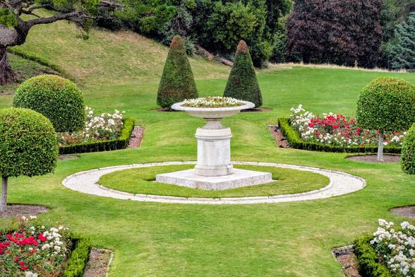A photograph showing a perfectly manicured garden with a central circular stone planter on a pedestal filled with white flowers. The planter is surrounded by a circular gravel path. On either side, there are symmetrically placed hedges and bushes of varying shapes, including two large conical-shaped topiary trees. Patches of red and white roses are visible in the foreground and the background.