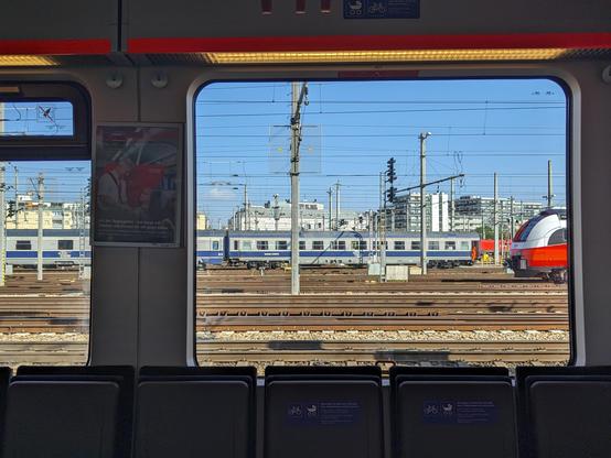 the window of an öbb talent S60. there's CFR sleeping wagons on another track. on the frontmost wagon there's a worker in an orange vest sitting in the open door