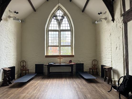 The inside of a small chapel. Underneath a large glass-in-lead window is a wooden table to either side of which a camp bed has been set up.