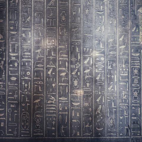 An ancient Egyptian stone tablet is covered in columns of engraved hieroglyphs. The carvings are stylised and intricate, depicting various symbols, animals, and human figures. The stone is dark, with lighter markings within the engraved lines. The tablet appears to be a formal inscription or a part of a larger monument, with the carvings arranged neatly in vertical lines.