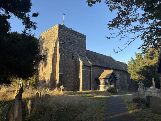 View of the church. The bell tower is large and square and surrounded by trees. The sky is a perfect blue