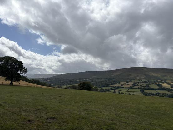 View over the Black Mountains. In the foreground is a green grassy field sloping downwards and in the middle and in the distance a big hill slopes upwards. The sky is cloudy with some blue patches.