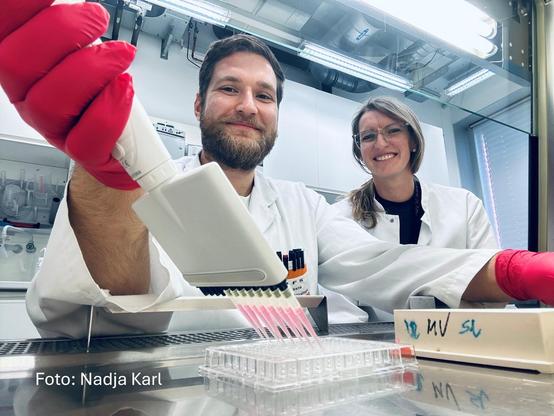 Ein Mann in weißem Kittel und mit roten Handschuhen pipettiert rosa Flüssigkeit unter einem Abzug im Labor. Neben ihm sitzt eine Frau in einem weißen Kittel.