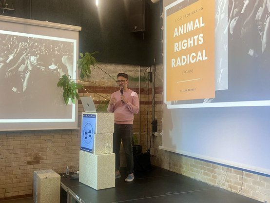 Jake speaks in front of an audience. The background is the fiest slide of his presentation, white letters over an orange block. The words "animal rights radical" are highlighted.