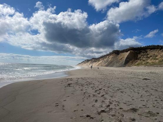 An ocean beach with sand dunes behind it and a partly cloudy sky above it.