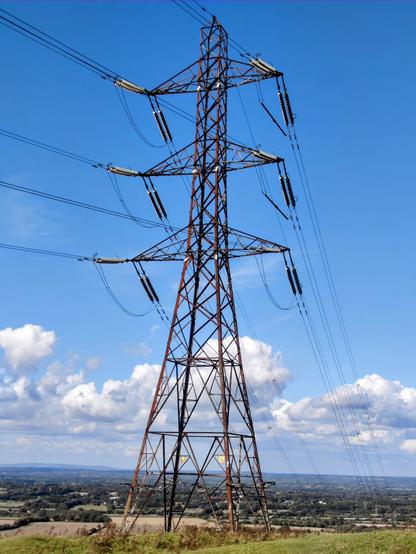 A large pylon with wires heading off up and down the hill in front of a long view of flat green countryside. A blue bright sky with scattered low white clouds.