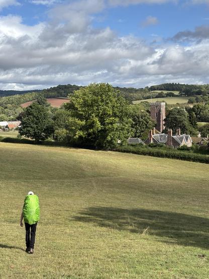 A grassy hill descending to Dore Abbey in the distance half hidden by trees. Halfway down the hill is my son with his big backpack covered by a bright green rain cover.