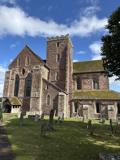A close up of Dore Abbey with the bell tower in the middle and the porch to the left. In the foreground are gravestones.