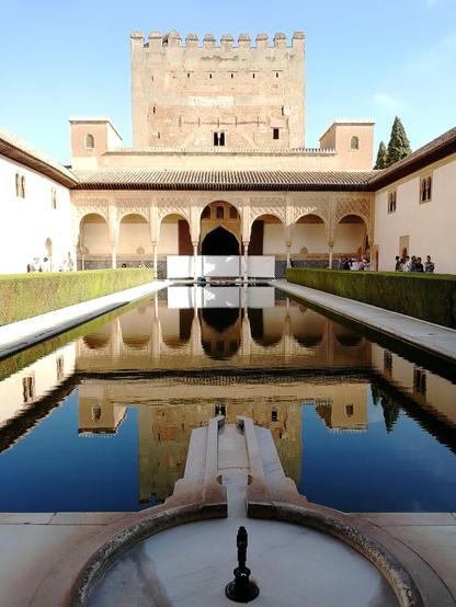 Bild aus dem Inneren der Alhambra: ein Gebäude spiegelt sich in einem See. Das Bild ist von einem direkt gegenüberliegenden Standpunkt aufgenommen, im Vordergrund befindet sich ein kleiner Brunnen.
