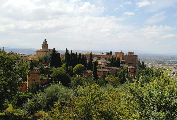 Man sieht die Alhambra von einem gegenüberliegenden Berg. Sie ist von Wald umgeben. Der Himmel ist blau.