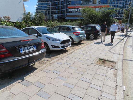 Street in a wealthier area of Tirana, Albania. The sidwalk is made of pavers. To the left are several cars parked on dirt next to the pavement. There are two tree wells that have trees that were sawn off low to the ground. Two men are walking along the sidewalk. Unseen to the right is bumper-to-bumper suburb commuter traffic.