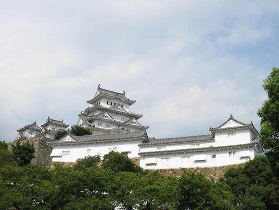 Image of Himeji Castle. Looking up from the southwest.
The castle tower (main keep) behind the long white walls, stone walls, and the green trees. There are a lot of clouds in the  sky.