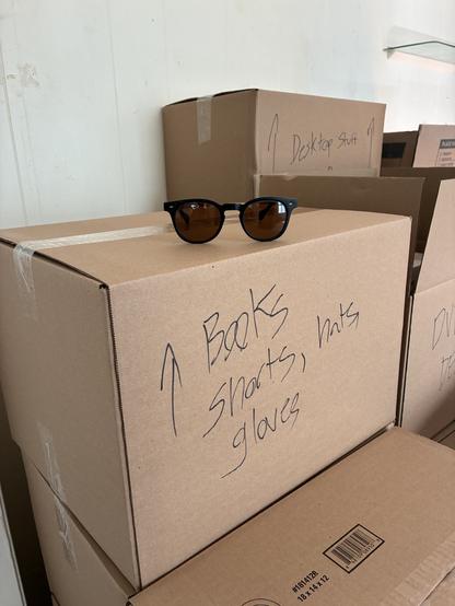 A pair of sunglasses atop a stack of boxes, the immediate one reading, "BOOKS, Shorts, hats, gloves," the one behind it, "Desktop stuff."
