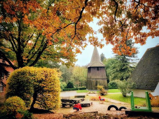A colourful autumn scene with golden and orange leaves on trees framing a rustic, wooden, conical-roofed building, possibly a church spire or windmill, in the centre. The foreground has a children's play area with a small red tractor toy and benches, surrounded by fallen leaves and lush green grass. The background shows more trees under a cloudy sky.