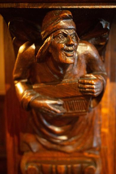 Color photo of a wooden carving of a figure wearing a head covering and holding a rectangular object, possibly a musical instrument.