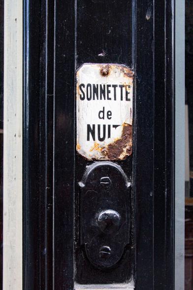 Color photo of a old, cracked enamel sign, mounted above a bell button. Th sign reads "Sonnette de Nuit"