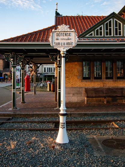 Color photo of a sign in an old fashioned design that says "Defense de circuler sur la voie". The sign is locatet on a rail track with a historic building in the background.