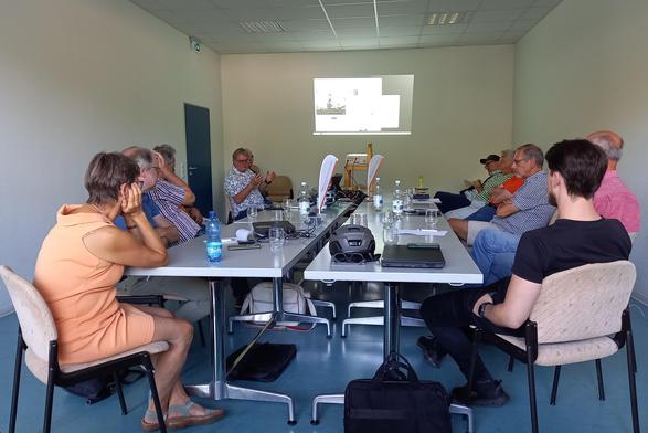 Blick in den Sitzungsraum im Bürgerhaus RSKN. Um eine lange, doppelte Tischreihe in der Mitte sitzt ein gutes Dutzend Teilnehmende. Sie blicken zum Referenten, der seitlich am Ende der Tischreihe sitzt. Auf die Wand hinter ihm wird mit einem Beamer eine Präsentationsfolie projiziert.