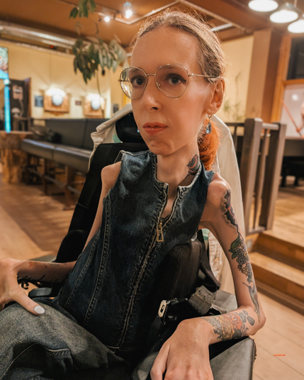 Ich sitze in einem warm beleuchteten Restaurant und trage einen ärmellosen Jeans-Jumpsuite mit Reißverschluss, eine runde goldene Brille, dezentes Make-up und Ohrringe. Sichtbar sind meine Tattoos an Unterarmen und Hals. Mein Blick ist ruhig und selbstbewusst in die Kamera gerichtet; im Hintergrund unscharf: Holzmöbel, Pflanzen und Deckenleuchten.