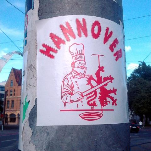 Aufkleber an einem Laternenmast in der Calenberger Neustadt in Hannover. Der typische Dönermann, der von vielen Dönerläden bekannt ist, schneidet Teile des Braunschweiger Löwen vom Spieß. Auf seiner Kochmütze ist das Logo von Hannover 96. Darüber steht das Wort »Hannover«.