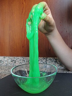 Eine Glasschale mit leuchtend grünem Slime. Eine Kinderhand hat einen größeren Teil davon festgehalten und nach oben gezogen. Der grüne Slime läuft langsam nach unten zurück.