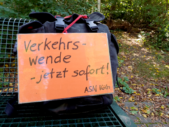 Auf einer Sitzbank steht ein Rucksack mit einem orangenen Schild: "Verkehrswende - jetzt sofort! (ASN Köln)"; im Hintergrund Büsche