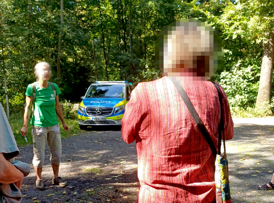zwei Frauen* stehen bei der Waldführung des BUND im Gremberger Wäldchen, im Hintergrund ein Polizeifahrzeug unter grünen Laubbäumen