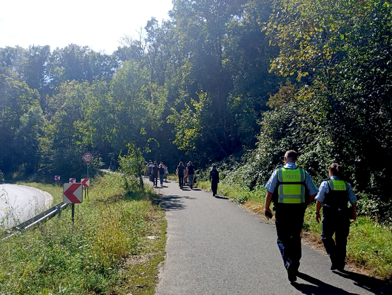 Am Rand einer Autobahn verläuft ein asphalierter Weg entlang des Waldrands, darauf gegen zwei Polizist*innen mit gelben Warnwesten; im Hintergrund Teilnehmer*innen des Waldspaziergangs im Gremberger Wäldchen
