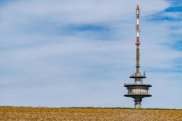 Photo of a 'Typenturm' standard radio tower peeking over a hill with a yellow stubble field.