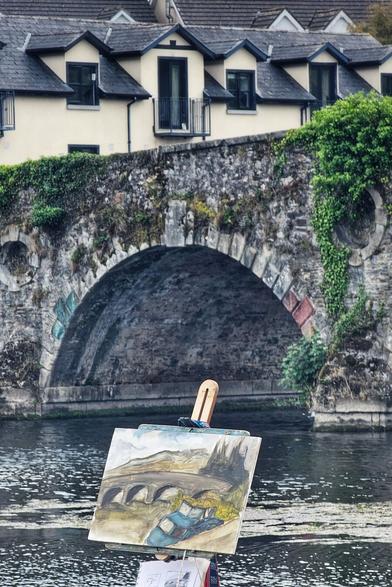 An easel with a watercolour painting of a stone bridge is set up on the bank of a river. The painting depicts the same stone bridge that is visible in the background. The bridge is overgrown with green ivy and has an archway that spans the river. In the background, a row of residential buildings with multiple dormer windows is visible above the bridge. The water is calm with a slight ripple.