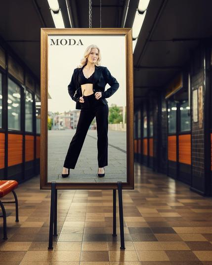 Framed fashion advertisement displayed in a subway setting. Features a woman in a black outfit with a blazer and pants, standing confidently with hands on hips. The word "MODA" is printed at the top left of the frame. Subway platform includes orange and tile elements with fluorescent lighting overhead.