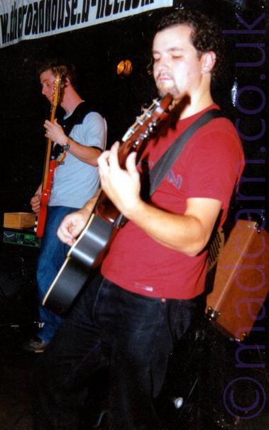 A view from the side of a man with short, curly dark hair, wearing a red t-shirt and a black pair of jeans, and playing a black acoustic guitar, looking down at his left hand as he stands facing off to the left of the frame.
An orange speaker is mounted at an angle behind him, while a taller man with short dark hair and wearing  a light and dark blue t-shirt is playing a red bass guitar off to the left of the frame.
Black walls and ceiling fill the rest of the frame.