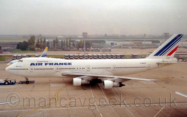 Side view of a very large, 4 engined jet airliner being towed from right to left by a low-slung white and blue tug attached to the nose-wheel by a long black pole.
The plane is almost entirely white, with large, blue, "Air France" titles on the upper forward fuselage, between the 2 passenger decks.
The blue registration "F-BPVP" is on the upper rear fuselage, and the last 2 letters are on the open nose-wheel doors.
The tail is white, with a series of 4 diagonal blue lines, getting thinner as they move rearwards, and a final, thick red stripe at the rear.
Grey concrete apron fills the foreground, with rows of buses parked side-by-side in the background, and white buildings and trees in the distance, slowly vanishing into the hazy grey sky.