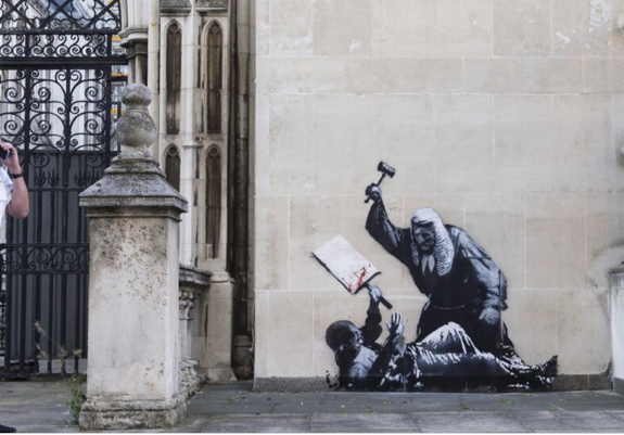This is a mural on a wall by the famous and elusive street artist named Banksy who documents with his work how men women and children are abused by government, powerful companies and companies. This drawing shows an British judge with a white wig, black robs and a gavel raised above his head attacking a young man of color on the ground who has a white placard raised in his left hand trying to shield himself from the blows from above.