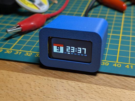 Ein 1,7″-LCD in einem blauen 3D gedruckten Gehäuse, welches die Uhrzeit und das Tagesdatum sowie den Wochentag anzeigt, auf einer grünen Arbeitsmatte auf einem Holztisch.