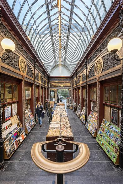 Symmetrical vertical view of the 19th century neo-Renaissance style covered passage Galerie Bortier in Brussels showcasing wrought iron, wood and marble works topped by an arched glass ceiling. On each side, old book stores. In the foreground a stylistic curved bronze hand rail leading down visitors to the book stores.
In 2024 this Galerie was transformed into a food hall and most of its book stores have now become little restaurants and cafes.