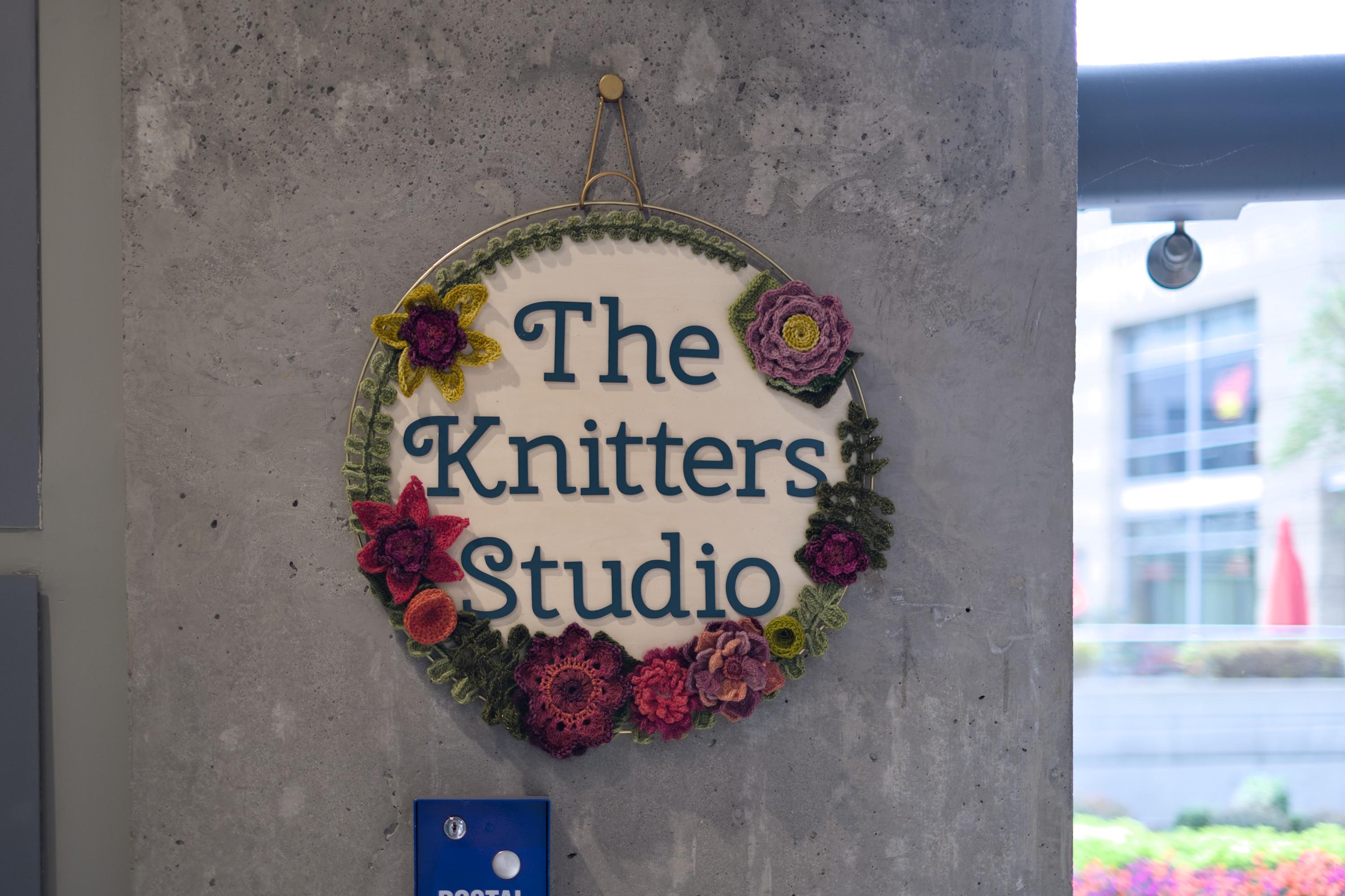 A round store sign saying "The Knitters Studio" with words in three rows, adorned with knitted flowers, hanging from a string on a concrete wall.