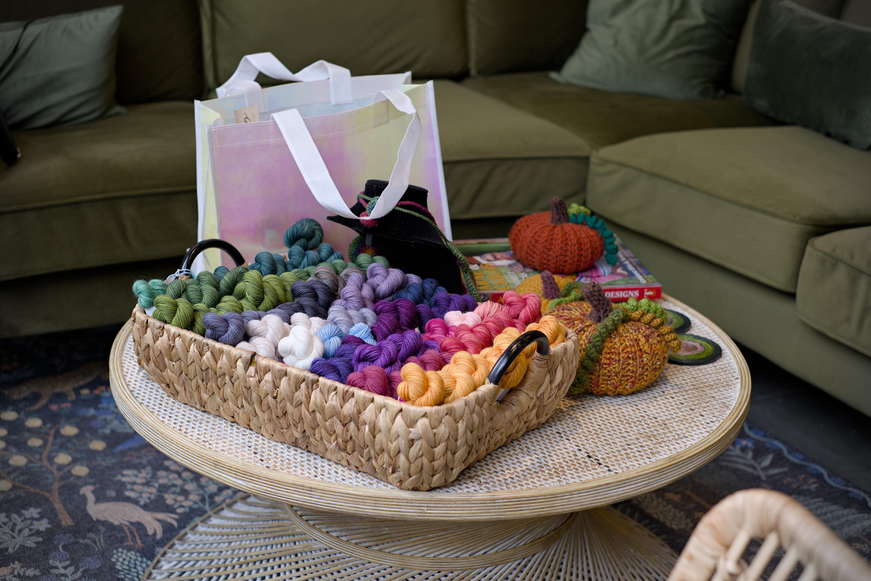A shallow basket of colorful skeins of yarn standing on top of a round low wooden table standing in front of an L-shaped green couch with decorative pillows.