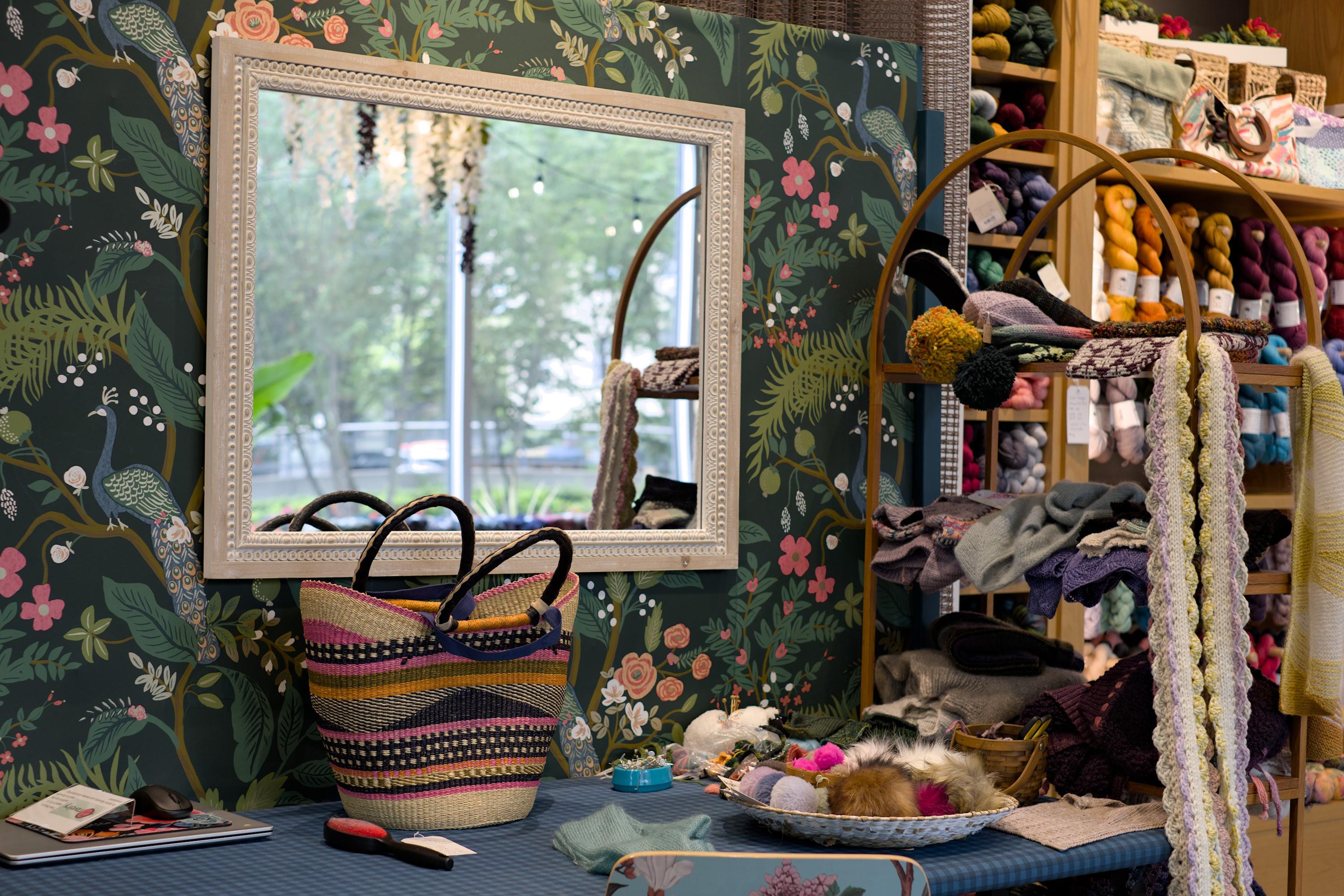 A carefully designed "working mess" table against a wall with a framed mirror on it. The wall is papered over with a flower motif. A colorful basket is standing on the table. A part of the yarn shelf is visible on the side of it.