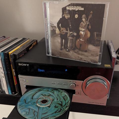 Una imagen que muestra un sistema de audio doméstico de la marca Sony. Sobre la unidad, un estuche de CD de pie. Es el álbum "In It For The Money" de Supergrass. La carátula muestra una fotografía desaturada o en tonos sepia de tres hombres: uno a la izquierda toca una guitarra acústica, el del centro mira hacia arriba con una expresión sorprendida, y el de la derecha toca un contrabajo. Los tres llevan gorras.

Debajo, el reproductor de CD, un modelo Sony CMT-SX7, tiene su bandeja abierta. Un disco con un diseño abstracto de trazos de pintura en tonos azules y turquesas está parcialmente fuera de la bandeja. El panel frontal del reproductor es plateado, con una pantalla que muestra la palabra "Open" en azul y un icono de Wi-Fi. A la derecha, se observa un gran dial de volumen plateado. A la izquierda del sistema de audio, se ve una pila vertical de otros estuches de CD o DVD.