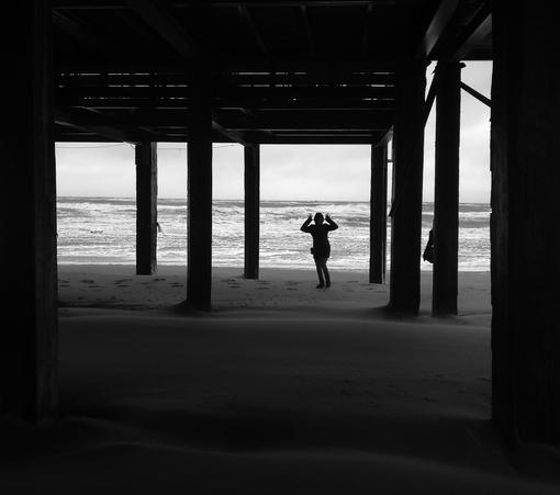 das bild zeigt eine schwarz-weiß-aufnahme von einer person, die unter einer holzkonstruktion zwischen hohen pfeilern am strand steht. die person ist als silhouette erkennbar und macht mit den händen eine art hasenohren-geste. der strand ist sandig und von wind gezeichnet, wie die wellen im meer im hintergrund. die säulen des steges rahmen den blick auf das stürmische meer. das gesamte bild wirkt düster und etwas melancholisch durch den kontrast zwischen der dunklen struktur des steges und dem hellen, aber unruhigen meer. der wind und die wellen lassen auf eine stürmische oder zumindest windige wetterlage schließen. die pose der figur, die den fotografen neckt, kontrastiert die szene.