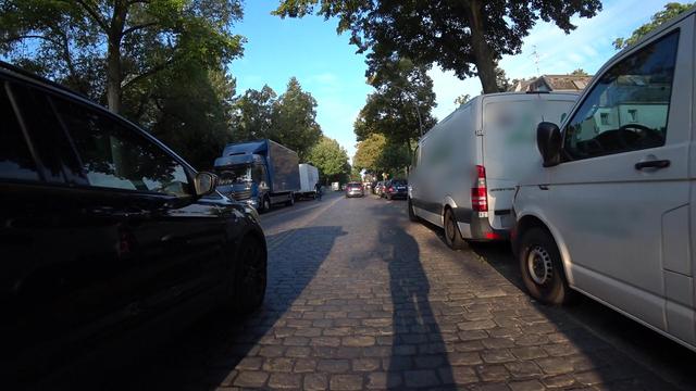Screenshot aus einer Cam. Blick auf eine Kopfsteinpflasterstrasse Rechts geparkte weiße Transporter mit geblurrten Firmenlogos, die über die Begrenzung der Parkbucht zum Teil in die Fahrbahn ragen, links der vordere Teil eines schwarzen PKW, der ca. 2m Abstand zu den geparkten Transportern hat. An den Schatten auf der Straße kann man erkennen, dass sich das Fahrrad mit der Cam ziemlich genau in der Mitte zwischen PKW und Transportern befindet, also links und rechts der Abstand geringer als 1m sein muss.