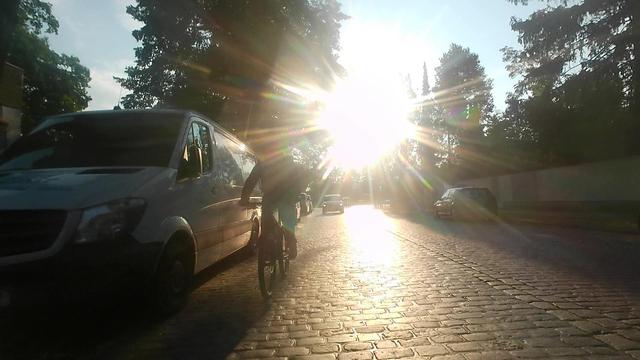 Screenshot aus der nach hinten gerichteten Cam. Kopfsteinpflasterstraße im "idyllischen" Gegenlicht. Links wieder ein weißer Transporter, der Radfahrer daneben hält mit Fahrradmitte gerade mal ca einen halben Meter Abstand vom Stehblech, bewegt sich also komplett in der Dooringzone.
