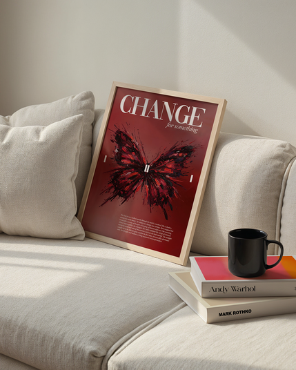 A wooden frame with a red butterfly poster rests against a beige sofa, beside two stacked art books by Andy Warhol and Mark Rothko with a black coffee mug on top, all bathed in soft natural light.